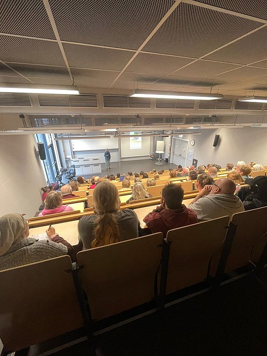Gesamtfoto des Hörsaals. Sitzende Menschen von hinten