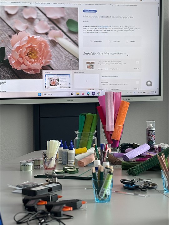 Foto vom Tisch auf dem verschiedene Bastelutensilien liegen. Im Hintergrund ist das Smartboard, das eine Kreppblume zeigt.