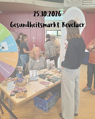 Glücksrad im Vordergrund. Im Hintergrund Monika, die an unserem Infotisch sitzt und sich mit einer Person unterhält, die vor dem Tisch steht